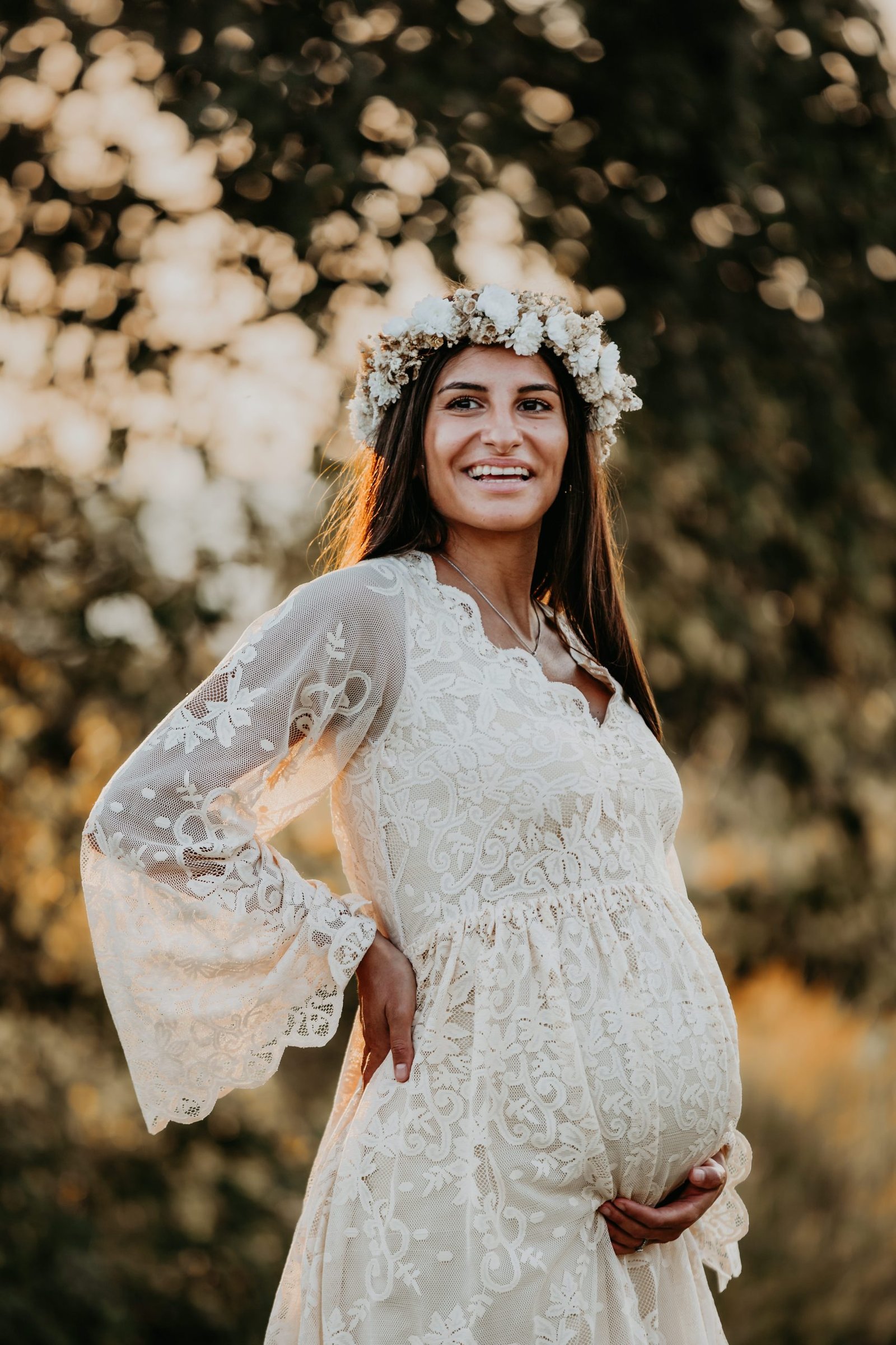 Gravid kvinna i spetsklänning som står i naturen i Skåne med blommor och mjukt kvällsljus.