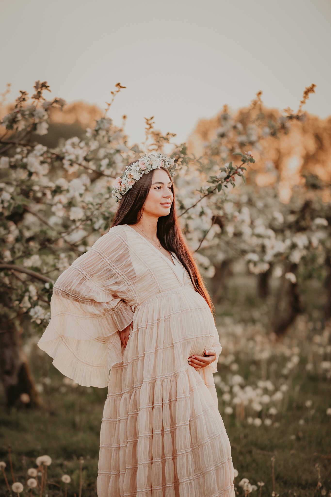 Gravid kvinna i ljus, flödande klänning och blomsterkrans som står i blommande natur i Skåne under mjukt kvällsljus.