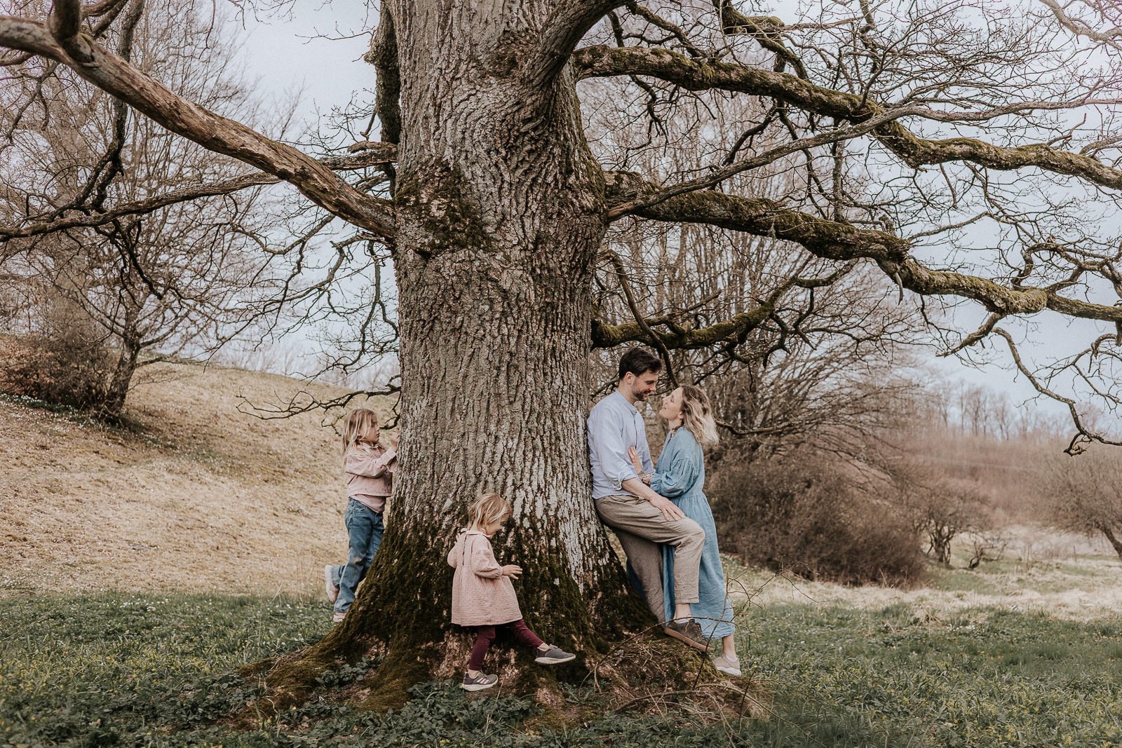 Naturlig familjefotografering i Skåne – lekfulla stunder i naturen Familj med två barn sitter och leker tillsammans på marken bland vita vårblommor under en naturlig familjefotografering i skogsmiljö i Skåne