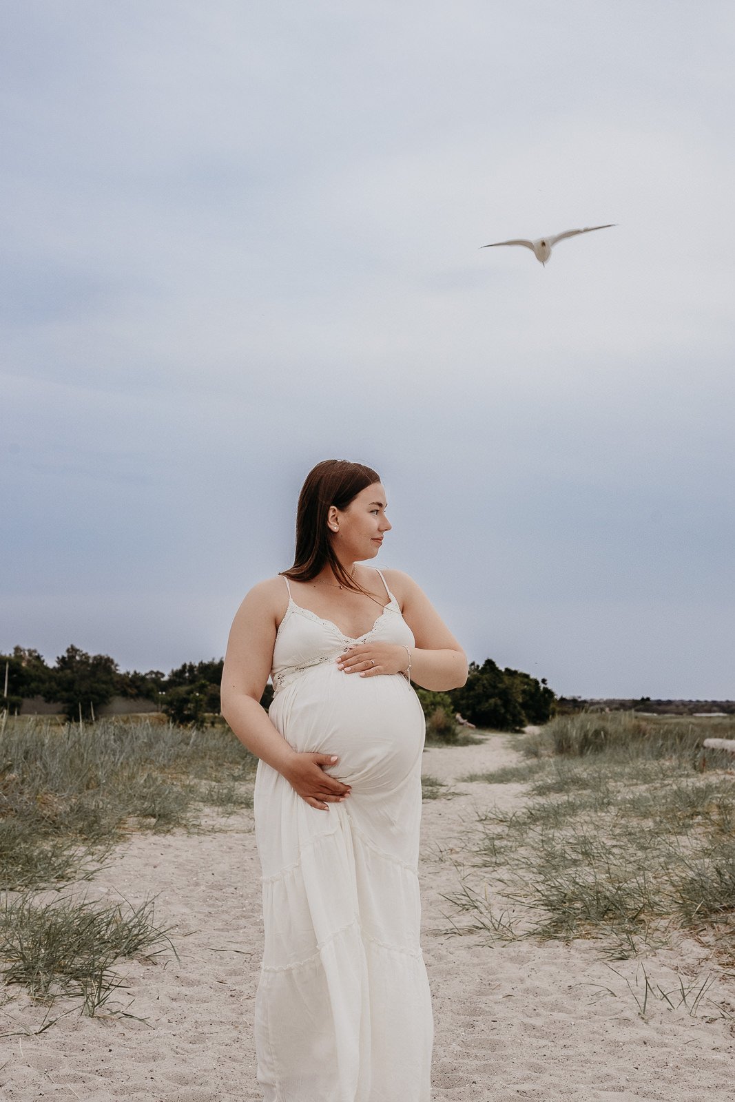 Gravid kvinna som står vid stranden i Malmö och håller sin mage i mjukt, naturligt ljus.